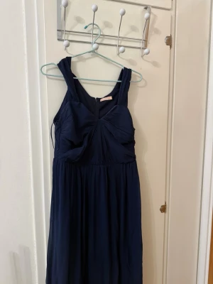 Balklänning  - Säljer en elegant marinblå långklänning med breda axelband och v-ringning. Klänningen har fina draperade detaljer över bysten och ett mjukt, flytande tyg som ger en snygg siluett. Perfekt för dig som vill ha en stilren och klassisk look.