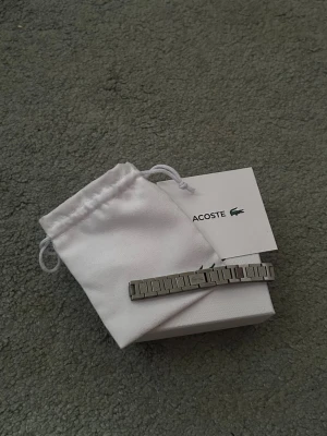 Silverfärgat armband från Lacoste - Snyggt armband från Lacoste i silverfärgad metall med stilren, länkad design. Kommer med originalpåse och ask med Lacoste-logga. Perfekt accessoar för dig som gillar minimalistisk och modern stil. Storlek kan skjusteras!