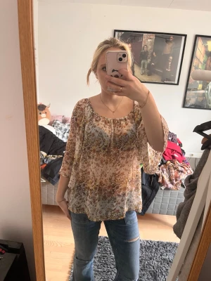 Blommig transparent blus med trekvartsärm - Säljer en luftig blommig blus i transparent material med beige, bruna och blå toner. Blusen har rundad halsringning och trekvartsärmar. Perfekt för dig som gillar en bohemisk och avslappnad stil.