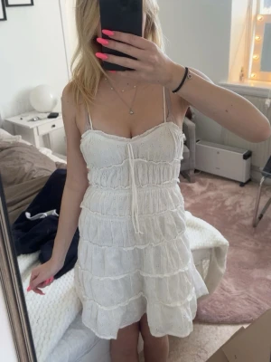 Vit somrig klänning med volanger - Söt vit klänning med tunna axelband och volangdetaljer i flera lager. Klänningen har broderade små blommor, knyt framtill och är i luftig bomull. Perfekt för varma dagar och ger en romantisk vibe.