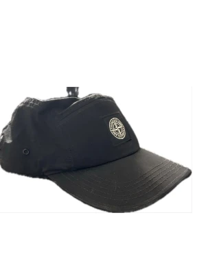 Svart keps från Stone Island - Snygg svart keps från Stone Island med klassisk logotyp framtill. Kepsen har böjd skärm, metallöljetter på sidorna och justerbart band bak med randig detalj. Tillverkad i lätt och slitstarkt material, perfekt för en streetwear-look.