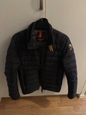 Svart pufferjacka från Parajumpers - Snygg svart pufferjacka från Parajumpers med hög krage, dragkedja framtill och två sidofickor med dragkedja. Jackan har det klassiska Parajumpers-emblemet på ärmen och en gul detalj vid halsen. Perfekt för kalla dagar.