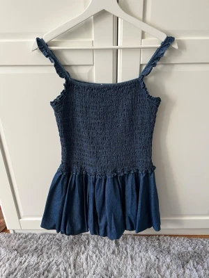 Mörkblå jeans illusions kort klänning med smock - Supersöt mörkblå kort klänning med smockad överdel och volangkant. Klänningen har breda axelband och en luftig kjol som ger en avslappnad vibe. Perfekt för varma dagar och enkel att styla med sneakers eller sandaler.