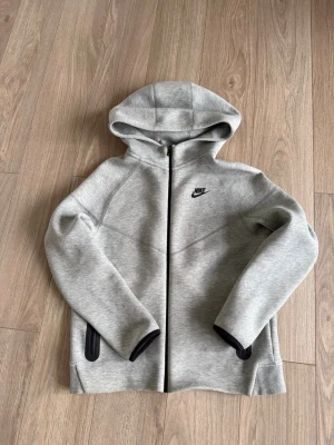 Grå Nike tech hoddie  - Snygg grå hoodie från Nike med svart logga på bröstet och hel dragkedja framtill. Hoodien har två svarta fickor med dragkedja och en stor huva. Tillverkad i mjukt och stretchigt material som känns riktigt bekvämt. Perfekt för en sportig och avslappnad stil. Lite sliten vid dragkedjan. Junior storlek 158-170.