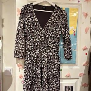 Brun omlottklänning med mönster - Snygg brun omlottklänning med vita och beige bladliknande mönster. Klänningen har v-ringning, trekvartsärm och knyt i midjan för en smickrande passform. Perfekt för dig som gillar unika prints och bekväma plagg.