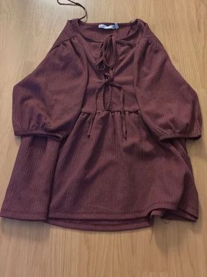 Vinröd blus med knyt och trekvartsärm - Snygg vinröd blus med struktur i tyget och två dekorativa knytband framtill. Blusen har trekvartsärmar med resår vid ärmslut och en avslappnad passform. Perfekt till jeans eller kjol för en trendig look.