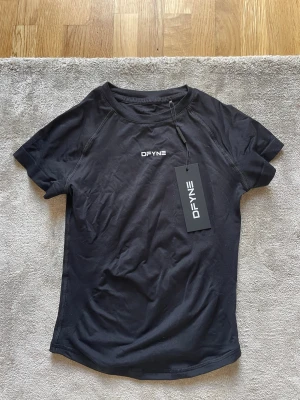 Svart träningströja från DFYNE - Snygg svart träningströja från DFYNE i modellen Impact Short Sleeve Top. Tillverkad i ett lätt och snabbtorkande material, perfekt för intensiva träningspass. T-shirten har korta ärmar och en diskret logga på bröstet.