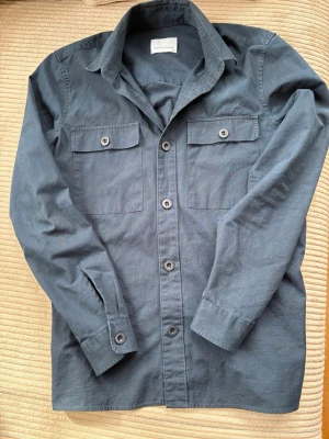 Mörkblå overshirt från Samsøe Samsøe - Stilren mörkblå overshirt från Samsøe Samsøe med två bröstfickor med lock och knappar. Klassisk krage och knäppning framtill. Perfekt lager på lager-plagg för en avslappnad look. Storlek S men passar även M.