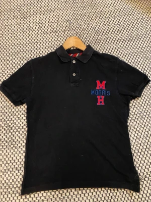 Svart pikétröja från Morris - Svart pikétröja från Morris med klassisk krage och två knappar. Broderad logga på bröstet i rött och blått med texten 'Morris' och bokstäverna M och H. Tillverkad i bomull och har korta ärmar för en clean och sportig vibe.