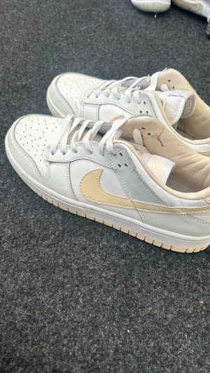 Nike Dunk Low vit/beige sneakers - Nike Dunk Low sneakers i vitt läder med beige swoosh och detaljer. Klassisk perforerad tåbox, vita skosnören och beige innersula. Yttersulan är beige och ger en snygg kontrast till den vita ovandelen. Perfekt för dig som gillar stilrena och tidlösa sneakers.