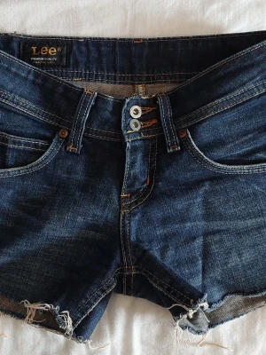 Mörkblå jeansshorts från Lee - Väldigt lowwaist så passar xxs-xs. Snygga mörkblå jeansshorts från Lee med råa kanter och dubbla knappar i midjan. Klassisk femficksmodell med kontrastsömmar och slitna detaljer för en avslappnad look. Perfekta för varma dagar.