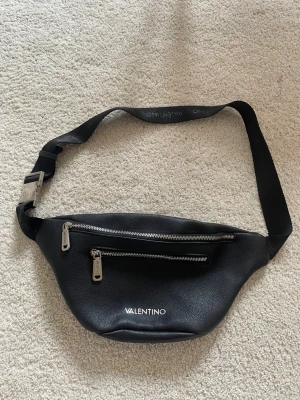 Svart handväska från Valentino - Snygg svart handväska från Valentino med justerbar axelrem i textil och detaljer i silverfärgad metall. Väskan har en dragkedja med Valentino-logga och är gjord i ett strukturerat konstläder som ger en lyxig känsla.