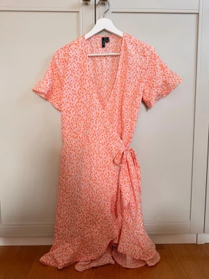 Omlottklänning i leopard från Vero Moda - Snygg omlottklänning från Vero Moda i vitt och orange med leopardmönster. Klänningen har korta ärmar, knyt i midjan och volangkant nedtill. 