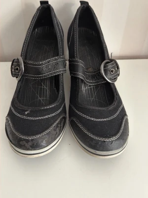 Svarta ballerinaskor med spänne - Snygga svarta ballerinaskor från Graceland med breda remmar och stora silverfärgade spännen. Skorna har vita kontrastsömmar och en rund tå. Materialet är en mix av textil och konstläder och konstläder har viss bortfall se bilder dock skorna är i bra skick. Cool och avslappnad look.