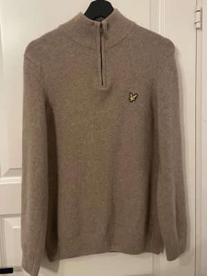 Beige halvzip-tröja från Lyle & Scott - Stickad beige tröja från Lyle & Scott med halvzip och hög krage. Tröjan har ett broderat gult örnmärke på bröstet och ribbade muddar vid ärmslut och nederkant. Perfekt för lager på lager och en clean look.