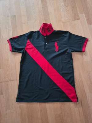 Svart och röd piké från Ralph Lauren - Snygg svart pikétröja från Ralph Lauren med röd diagonal rand och röd logga på bröstet. Kragen och ärmsluten är röda, och tröjan har knappslå framtill. 