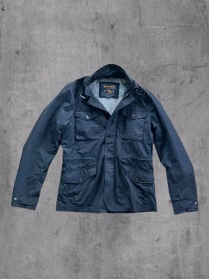 Marinblå vindjacka från Woolrich - Snygg marinblå vindjacka från Woolrich med fyra stora fickor framtill och hög krage. Jackan har knapp- och dragkedjestängning samt justerbara muddar. Tillverkad i ett lätt, vattenavvisande material som passar perfekt för blåsiga dagar. Liten defekt på ena fickan syns på bild 