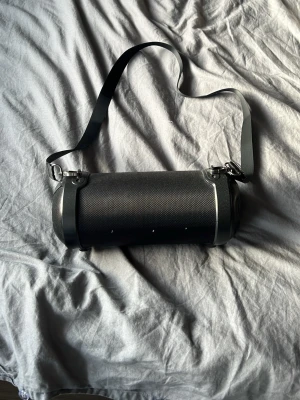 Svart cylinderformad bärbar högtalare - Snygg svart bärbar högtalare med cylinderformad design och axelrem för enkel transport. Högtalaren har metalliska detaljer på sidorna och ett robust galler i plast eller metall runt om. Perfekt för att ta med sig musiken överallt.