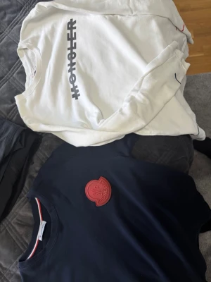 Moncler tröjor i vitt och marinblått - Två snygga tröjor från Moncler, en vit med stor logotyp i svart på bröstet och en marinblå med röd Moncler-patch. Båda har rund hals och klassisk passform. Perfekta för dig som gillar stilrena och exklusiva plagg.
