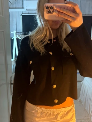 Svart croppad kofta med guldknappar - Trendig svart croppad kofta med långa ärmar och snygga guldknappar framtill. Två bröstfickor med dekorativa knappar ger en cool touch. Perfekt att styla till både jeans och kjol för en modern look.
