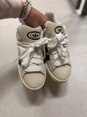 Adidas Campus vita och svarta sneakers - Snygga Adidas Campus sneakers i vitt mocka med svarta detaljer och klassiska tre ränder på sidan. Skorna har tjocka vita skosnören, gummisula och Adidas-logga på plösen. Perfekta för dig som gillar retrostil och streetwear.