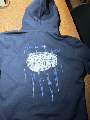 Mörkblå Bvaldi hoodie - Cool mörkblå hoodie från Svaldi med stor grafisk print på ryggen av en klocka och texten 'Who cares I'm already late' i blått och vitt. Hoodien har huva och är tillverkad i mjukt material, perfekt för en avslappnad streetwear-look.