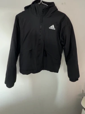 Svart vindjacka med huva från Adidas - Svart vindjacka från Adidas med vit logga på bröstet och tre vita ränder på ryggen. Jackan har huva, dragkedja framtill och två snedställda fickor. Perfekt för blåsiga dagar och sportig stil.