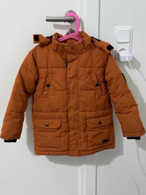 Rostbrun parkas med huva - Snygg rostbrun parkas med huva och fyra fickor framtill. Jackan har svarta detaljer vid ärmsluten och knappar på fickorna. Perfekt för kyliga dagar och har en modern, quiltad design. Stängs med dragkedja och tryckknappar.