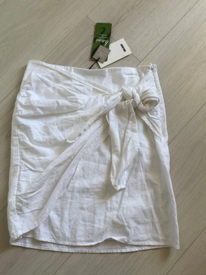 Vit omlottkjol i linne med knyt - Snygg vit omlottkjol  i luftig linneblandning. Kjolen har en trendig knytning i midjan och en kort längd som passar perfekt till varma dagar. Enkel och stilren design som är lätt att matcha med olika toppar.