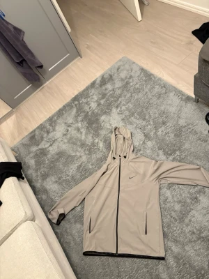 Beige vindjacka med huva från Nike - Snygg beige vindjacka från Nike med hel dragkedja framtill och huva. Jackan har svarta detaljer vid dragkedjor och muddar samt två sidofickor. Perfekt för blåsiga dagar och enkel att matcha med olika outfits.