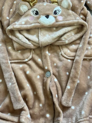 Brun prickig morgonrock med huva - Supersöt brun morgonrock i mjuk fleece med vita prickar och huva som har ett broderat djuransikte, öron och guldfärgade horn. Perfekt för mysiga hemmakvällar! Stängs med knappar framtill och har två fickor.