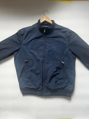 Marinblå bomberjacka från Polo Ralph Lauren - Snygg marinblå bomberjacka från Polo Ralph Lauren med dragkedja framtill, två snedställda fickor med dragkedja och ribbade muddar vid ärmslut och nederkant. Diskret logga broderad på bröstet. Tillverkad i ett lätt, slitstarkt material.