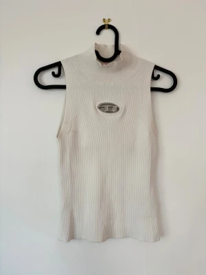 Diesel Vit ribbad ärmlös polotopp  - Trendig vit ribbad polotopp med hög krage och ärmlös design. Toppen har en unik metall-detalj framtill som ger en cool edge. Perfekt att styla med jeans eller kjol för en modern look. Nypris 2500 sek. Prislapp finns kvar.