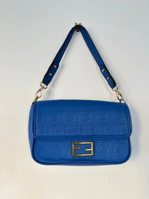 Blå handväska fendi vintage  - Snygg blå handväska med präglat FF-mönster över hela väskan och guldfärgat spänne framtill. Väskan har en justerbar axelrem i matchande blått och silverfärgade metalldetaljer. Klassisk rektangulär form och stilren design.