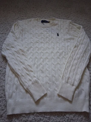 Vit kabelstickad tröja Polo Ralph Lauren - Kabelstickad vit tröja från Polo Ralph Lauren med klassisk rund halsringning och svart broderad logga på bröstet. Tröjan har ribbade muddar vid ärmslut och nederkant, perfekt för dig som gillar stilren och tidlös design. Passar mer som S.