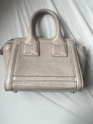 Beige handväska med krokopräglat mönster - Snygg beige handväska i konstläder med krokopräglat mönster och två stadiga handtag. Väskan har en boxig form och dragkedja upptill. Perfekt för dig som gillar stilrena accessoarer med lite edge.