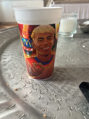 McDonald's plastmugg med fotbollstryck - Cool plastmugg från McDonald's med färgglatt motiv av en fotbollsspelare och texten 'Winner Yama'. Muggen har en vit kant upptill och är dekorerad i rött, gult och blått med tecknad stil. Perfekt för dig som gillar sportiga samlarprylar.