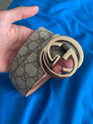 bälte med guldspänne - Snyggt bälte med klassiskt monogrammönster i beige och grått. Bältet har ett stort, rundat guldspänne. Perfekt accessoar för att lyfta vilken outfit som helst.