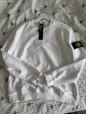 Vit sweatshirt från Stone Island - Snygg vit sweatshirt från Stone Island med klassisk rund hals och ribbade muddar. Tröjan har den ikoniska svarta logotypen med kompass på vänster ärm. Tillverkad i mjuk bomull för en skön och avslappnad look.