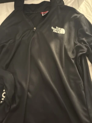 Svart långärmad tröja The North Face - Svart långärmad sporttröja från The North Face med dragkedja vid halsen. Logga tryckt på bröstet och längs ena ärmen i vitt. Tillverkad i ett glansigt, snabbtorkande material som passar perfekt till träning eller utomhusaktiviteter.