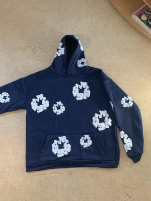 L. Denim tears mörkblå hoodie - Äkta denim tears har aldrig använt fick den som present av min store bror passar inte min stil har casual/classic stil helt ny inga fläckar eller hål hellt ny ingen prislapp pågrund av det är en present Snygg mörkblå hoodie med stora vita blommönster över hela tröjan. Hoodien har en klassisk huva och en stor magficka framtill. Perfekt för dig som gillar streetwear och vill sticka ut med ett unikt mönster. Materialet känns mjukt och bekvämt.