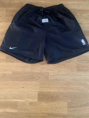 Svarta meshshorts Air Fear of God - Säljer ett par svarta meshshorts från Air Fear of God x Nike med NBA-logga och Nike Swoosh. Shortsen har snörning i midjan och är tillverkade i ett ventilerande polyestermaterial. Perfekta för basket eller chill.