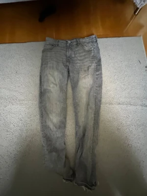Snygga gråa jeans - Säljer ett par grå jeans med tvättad look och slitningar. Jeansen har raka ben och råa, fransiga benslut för en avslappnad vibe. Klassisk femficksmodell och knappgylf. Perfekta för dig som gillar en edgy stil.