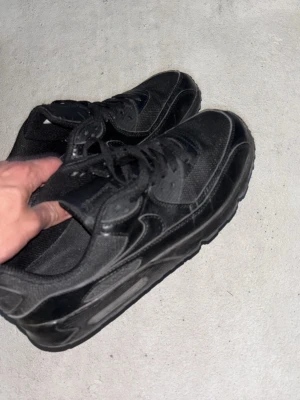 Svarta Nike Air Max sneakers - Säljer ett par helsvarta Nike Air Max sneakers med klassisk chunky sula. Skorna har ovandel i både mesh och läder, samt svarta skosnören. Swoosh-loggan syns på sidan och designen är tidlös och clean.
