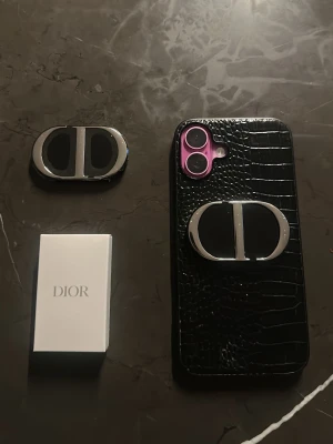 Dior svart mobilskal med spegel - Snyggt svart mobilskal från Dior med krokodilstruktur och silverfärgad logga. Skalet har en inbyggd spegel som fälls ut på baksidan, perfekt för snabba koller. Passar till dig som vill ha en lyxig och trendig look på mobilen.