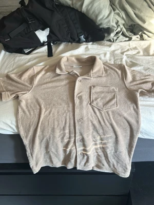 Beige frottéskjorta med kort ärm - Säljer en beige skjorta i mjuk frotté med korta ärmar och klassisk krage. Skjortan har knappar framtill och en bröstficka för chill stil. Perfekt för dig som gillar avslappnade och trendiga plagg.