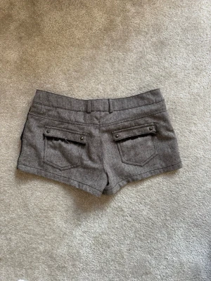 Gråa shorts med snygga fickdetaljer - As snygga grå shorts med två bakfickor med knappdetaljer och bälteshällor. Passar verkligen till allt och kramar om rumpan exakt som man vill. Perfekta nu till sommaren till utekvällar med ett par boots till och en snygg top. Står 34 men passar mig som har 36. 