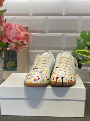 Maison Margiela sneakers med färgstänk - Unika vita sneakers från Maison Margiela med färgstänk i rött, gult, svart, turkos och grönt. Skorna har klassisk låg siluett, vita snören och gummisula. Tillverkade i läder med en cool konstnärlig vibe som sticker ut.