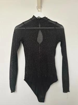 Svart glittrig mesh-body med lång ärm - Bershka  - Snygg svart body i mesh med glittereffekt och lång ärm. Hög hals med liten volangkant och en dekorativ öppning fram och bak. Tunt, stretchigt material som ger en vacker look. Inköpt på Bershka. Använd men i fint skick. Storlek M. 
