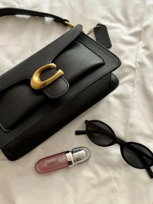Svart Coach Tabby 26 handväska  - Snygg svart handväska från Coach i  pebble-läder med guldfärgat C-spänne framtill. Väskan har en stilren, rektangulär form och justerbar axelrem. Perfekt accessoar för att lyfta din outfit med en lyxig touch.